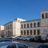 Oltre le auto si staglia il bianco prospetto della Corridoni, il cui aspetto imperioso rimanda subito alla sua epoca di costruzione. Fu infatti innalzata durante il fascismo, nel vivo dei lavori di risanamento di Bari vecchia